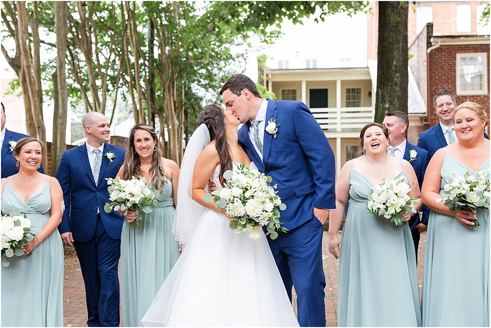 bride and groom share kiss while wedding party flanks them for a portrait before classic southern wedding