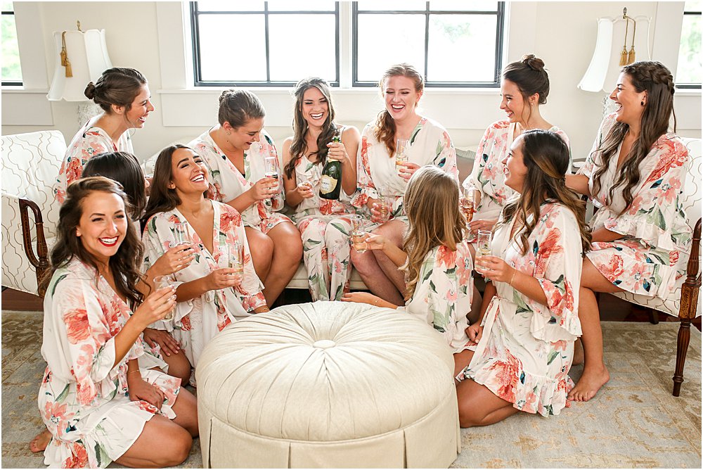 Bridal party in floral robes gather around the bride who is popping champagne during her stress free wedding day.