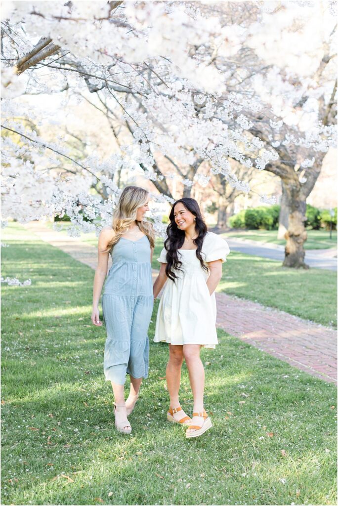 Ellen and Keira walk together, holding hands beneath cherry blossoms during their joint senior portrait session in Richmond va