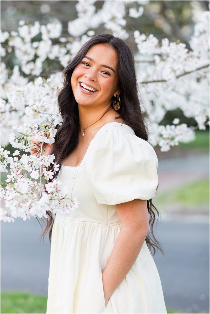 Keira smiles widely as she fiddles with a tree branch surrounded by pastel cherry blossoms during the spring senior portrait session