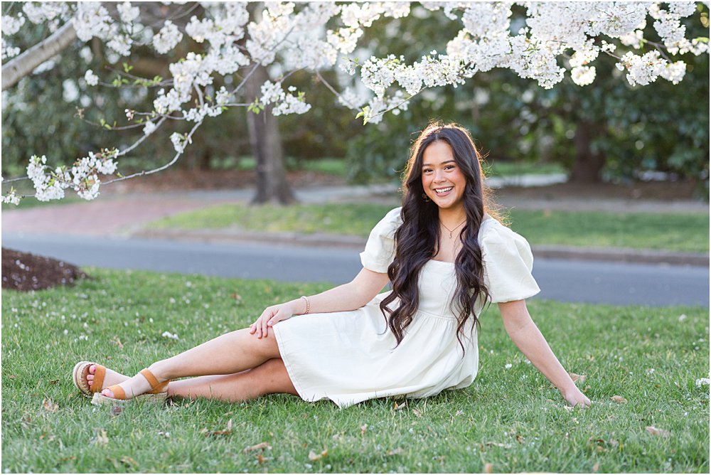 Keira stretches out on the green grass, smiling, beneath the cherry blossoms during her spring senior portrait session in RVA