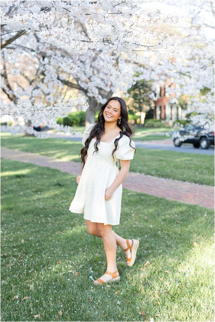 Keira stands beneath the cherry blossoms on green lawn during Spring senior portrait session in Richmond, VA