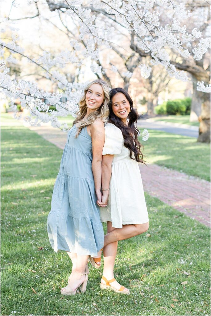 Ellen and Keira stand back to back , clasping hands beneath cherry blossoms during their spring senior portrait session in Richmond, VA
