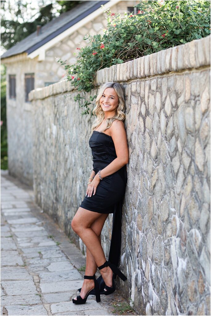High school senior poses for a photo in black mini dress and heels in front of stone wall during bright and bubbly senior portrait session in Richmond, VA