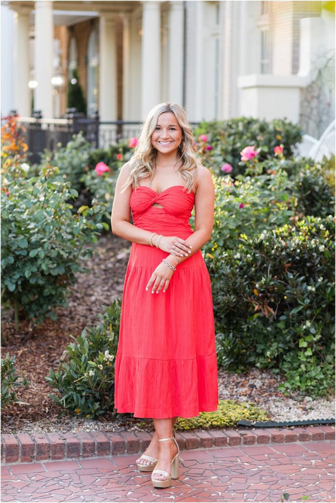 High school senior, wearing read strapless dress, poses for a photo on Monument Avenue in front of greenery and rose bushes during bright and bubbly senior portrait session in Richmond, VA