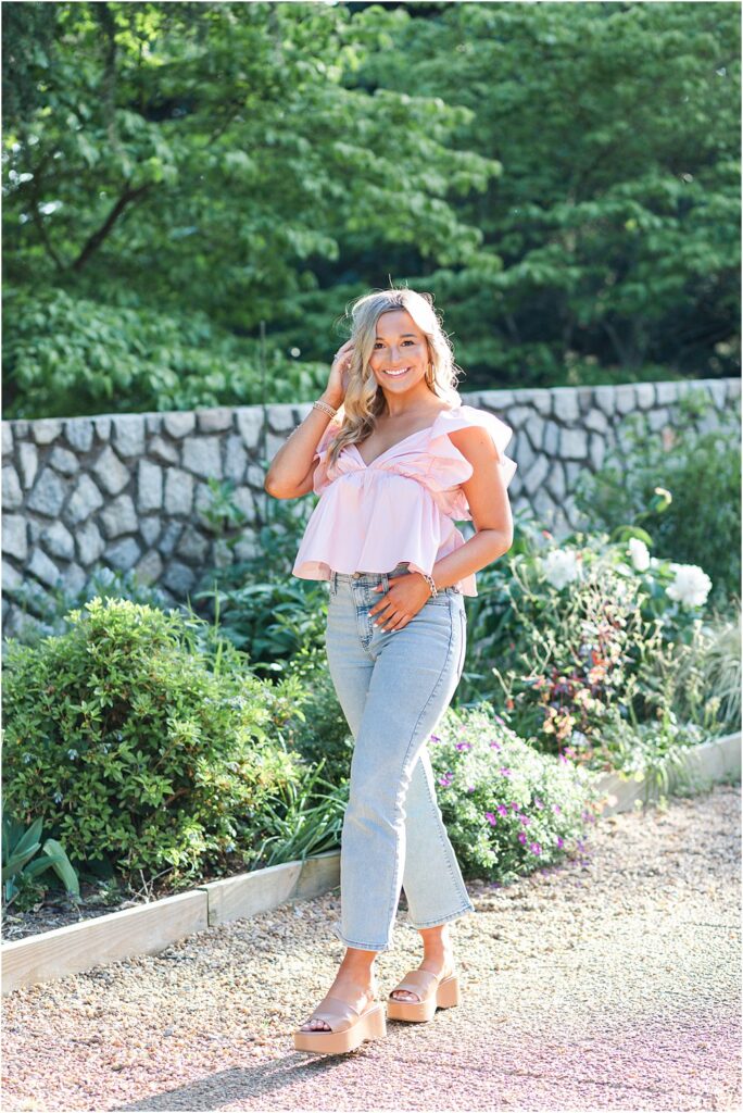High school senior, wearing pale blue denim jeans and blush puffed butterfly sleeve blouse, poses for a photo in Maymont Park during bright and bubbly senior portrait session in Richmond, VA