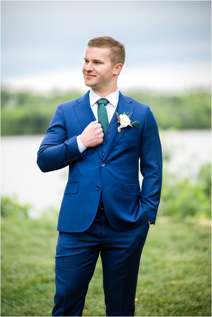 The groom, John, stands in a blue suit front of the river at Upper Shirley.