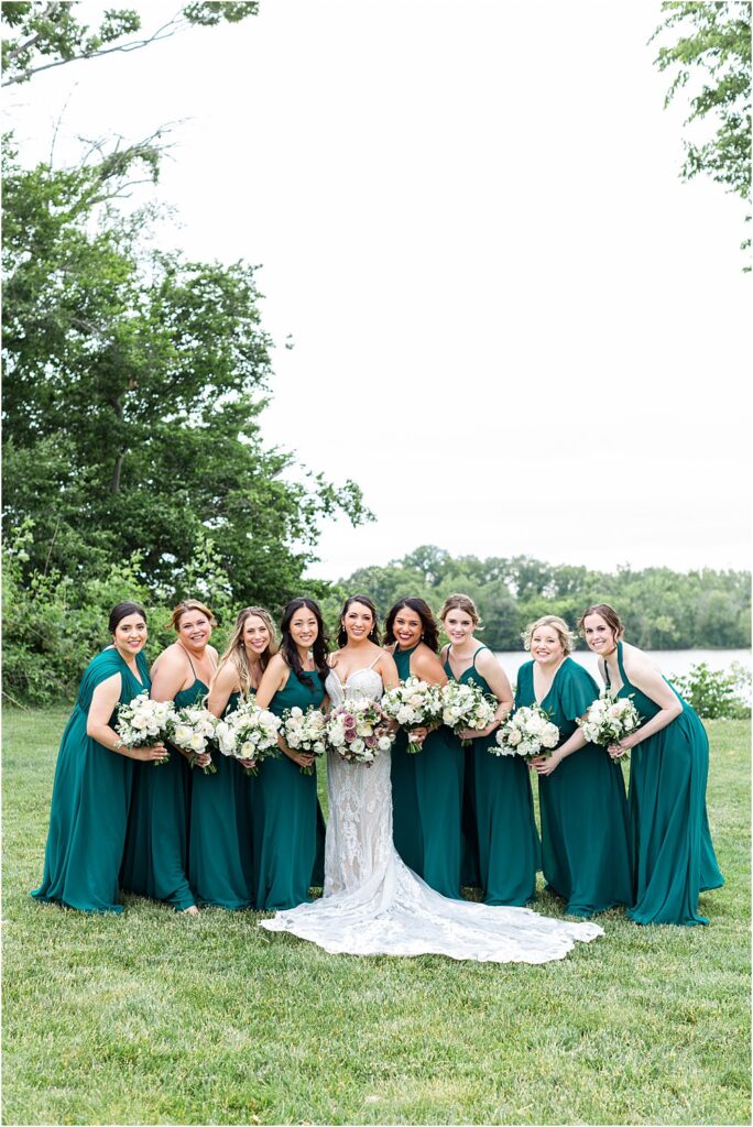 Bridal party, dressed in long emerald green dresses, surround the bride while holding bouquets on the lawn overlooking the James River at Upper Shirley.