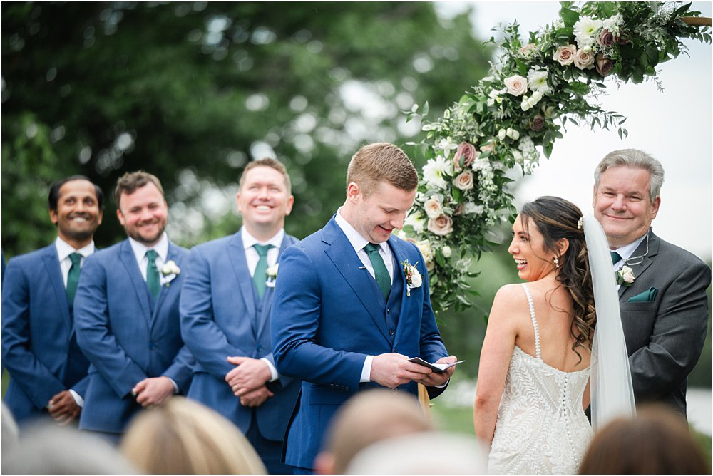 Wedding guests, groomsmen, and Stephanie laugh as John reads his wedding vows during modern romantic wedding ceremony.