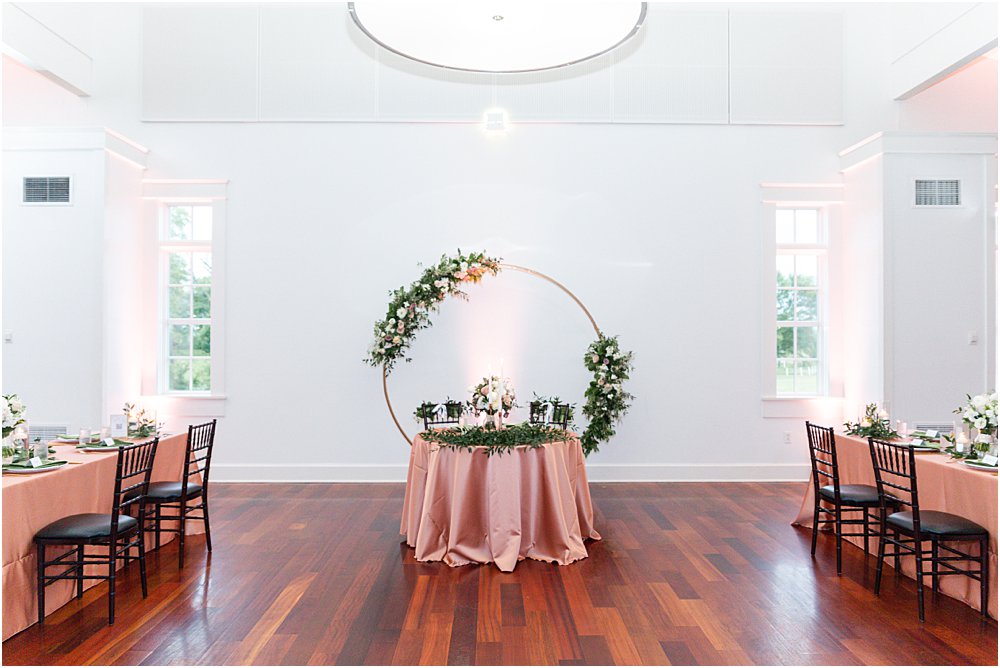 Reception setup in the ballroom at Upper Shirley Vineyards. Sweetheart table, draped in blush silk tablecloth, adorned with greenery. Behind it sits a circular structure adorned with flowers for the couple's modern romantic wedding in Charles City, VA