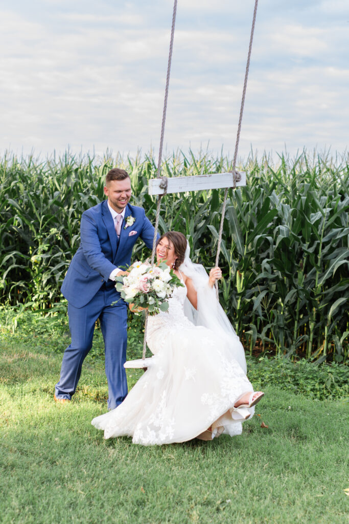 groom pushes bride on a swing in front of corn field during romantic barn wedding reception
