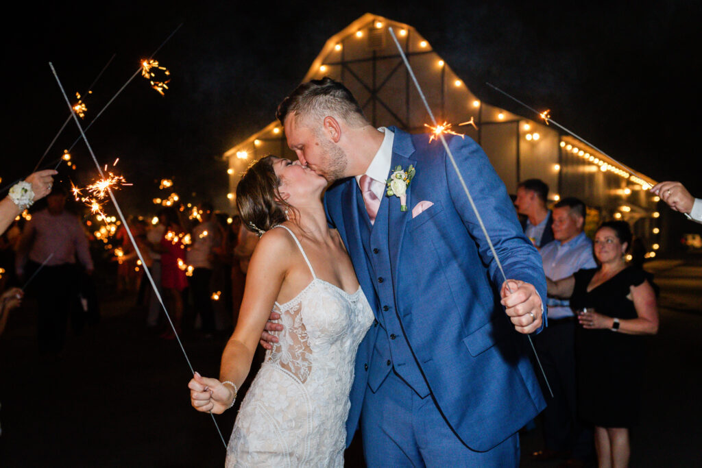bride and groom kiss while friends and family wave sparklers during grand exit at the end of romantic barn wedding day