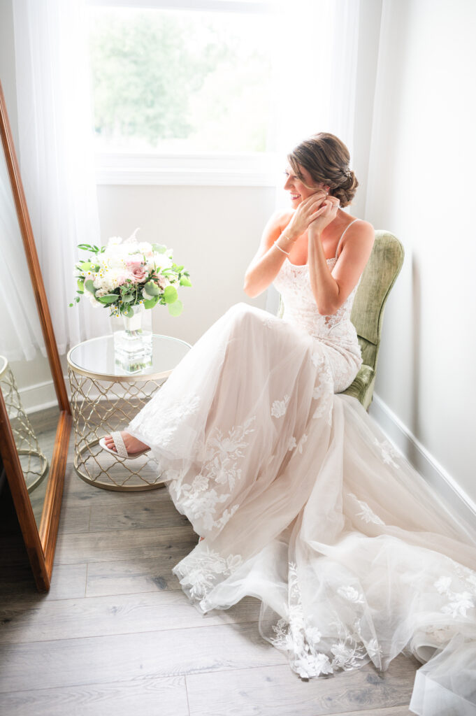 bride, seated by window, is encased by soft lighting as she dons her earrings.