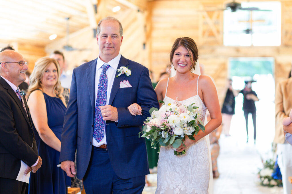 The bride grins from ear to ear as her father walks her down the aisle during romantic barn wedding ceremony in King William, VA