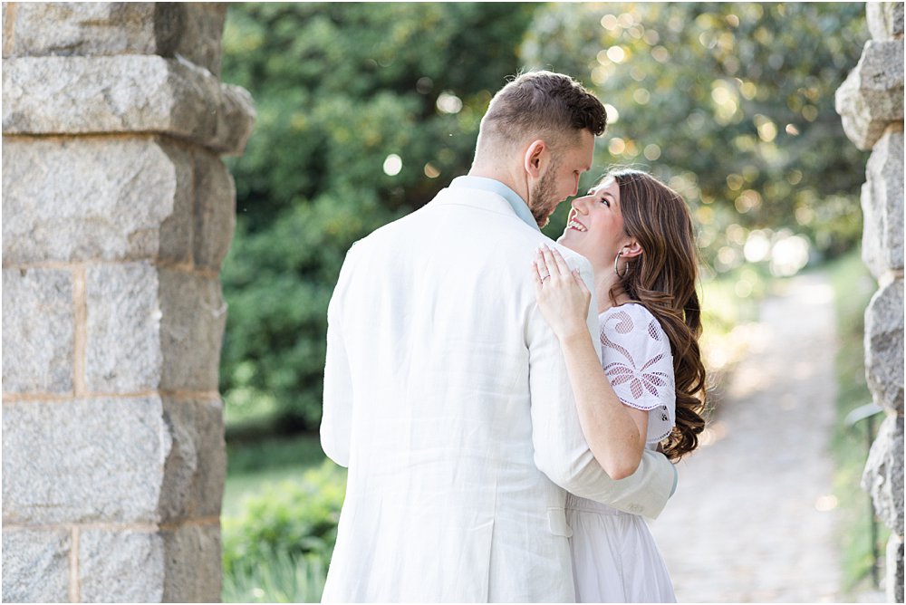 The couple, smiling and nose to nose, prepare to kiss beneath a stone arch at Maymont during their effortlessly chic engagement session.