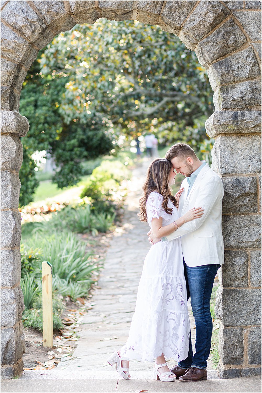 The couple, eyes closed and forehead to forehead, stand beneath a stone arch at Maymont Park during their effortlessly chic engagement session.