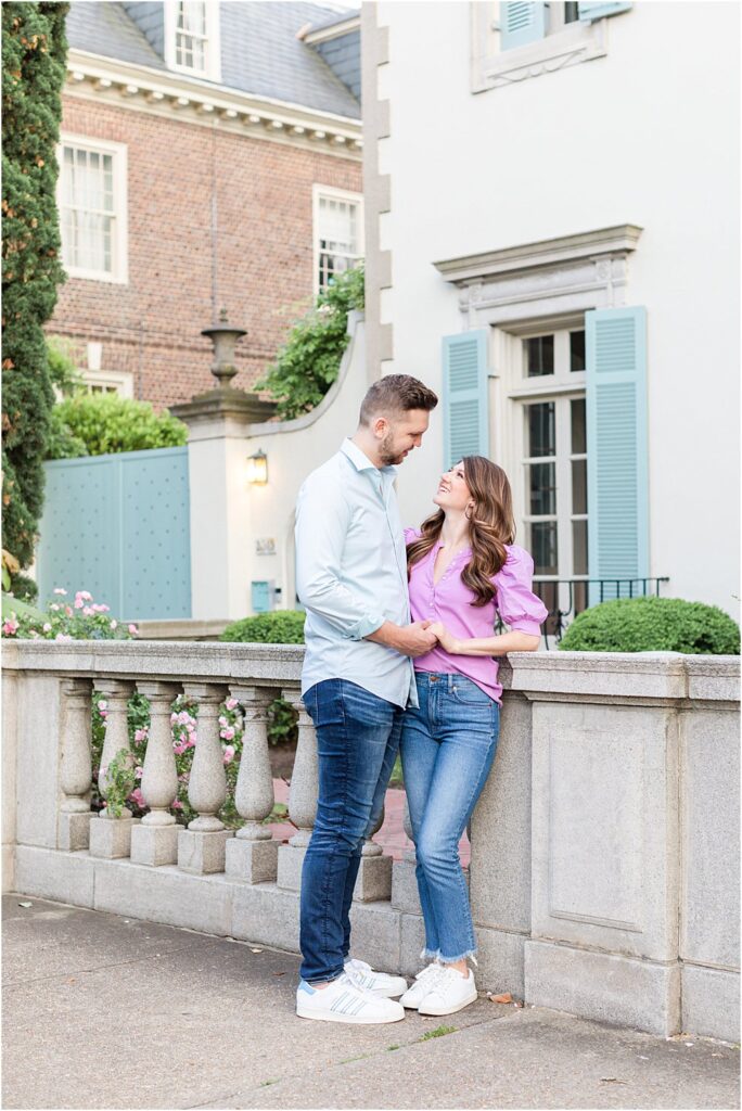 The couple stops to lean against a balustrade fence and gaze lovingly at one another while holding hands during their effortlessly chic engagement session in RVA