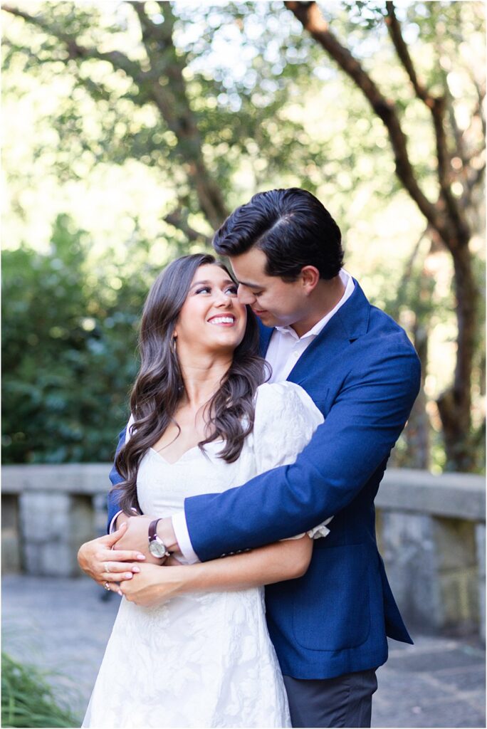 Raphael, stands behind Gabriella. He wraps his arms around her and snuggles into the soft sweetness of her hair. She smiles up at him during classic engagement session at Maymont Park in Richmond, VA.