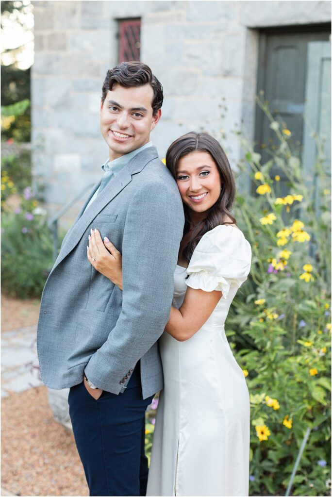 Gabriella, standing behind Raphael, rests her head against his shoulder as the pair smile directly at the camera during classic engagement session at Maymont Park in Richmond, VA.